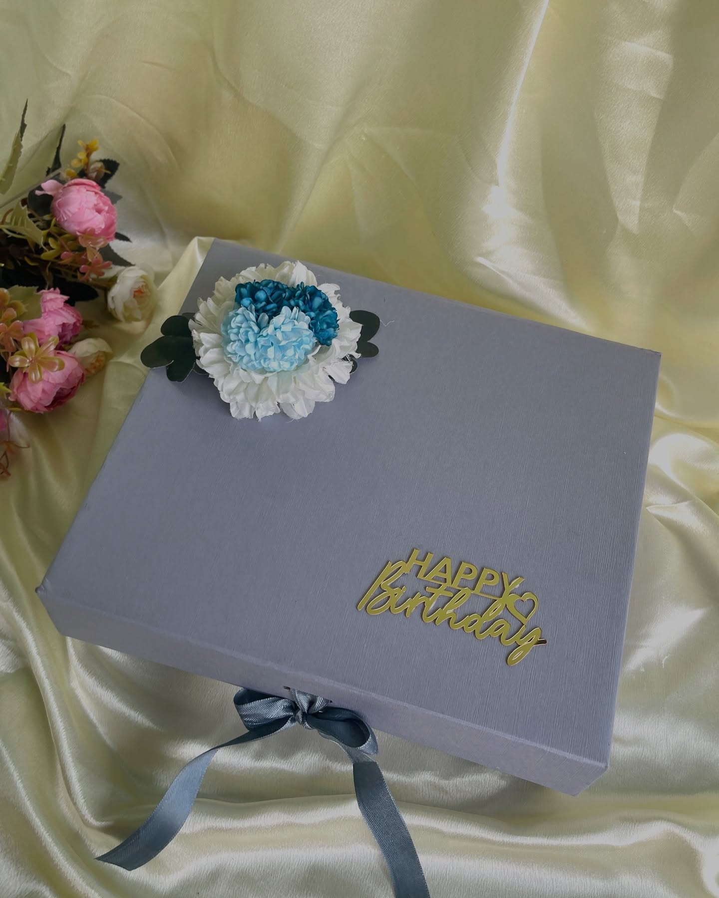 A sophisticated slate-grey gift box adorned with blue and white blooms and a gold Happy Birthday topper.