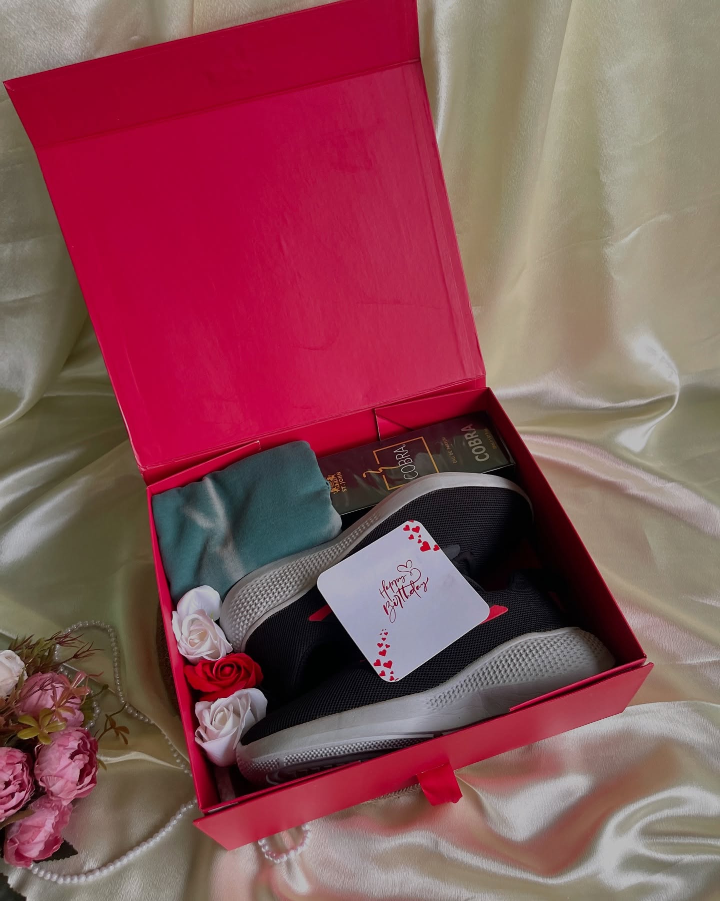 A bold pink gift box featuring sneakers, St. John Cobra cologne, a soft tee, and a handwritten birthday tag.