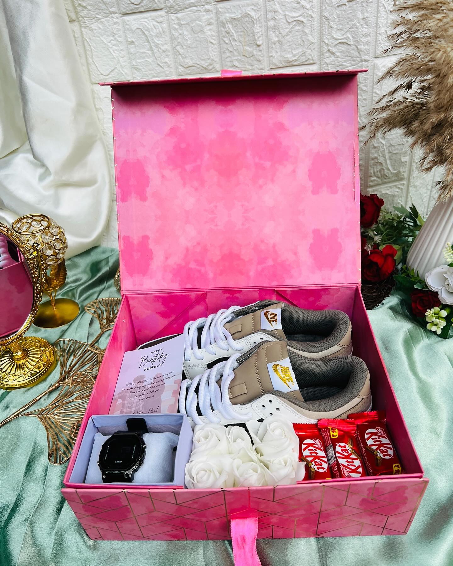 A pink birthday box with Nike sneakers, a black Casio watch, foam roses, KitKat bars and a message card.