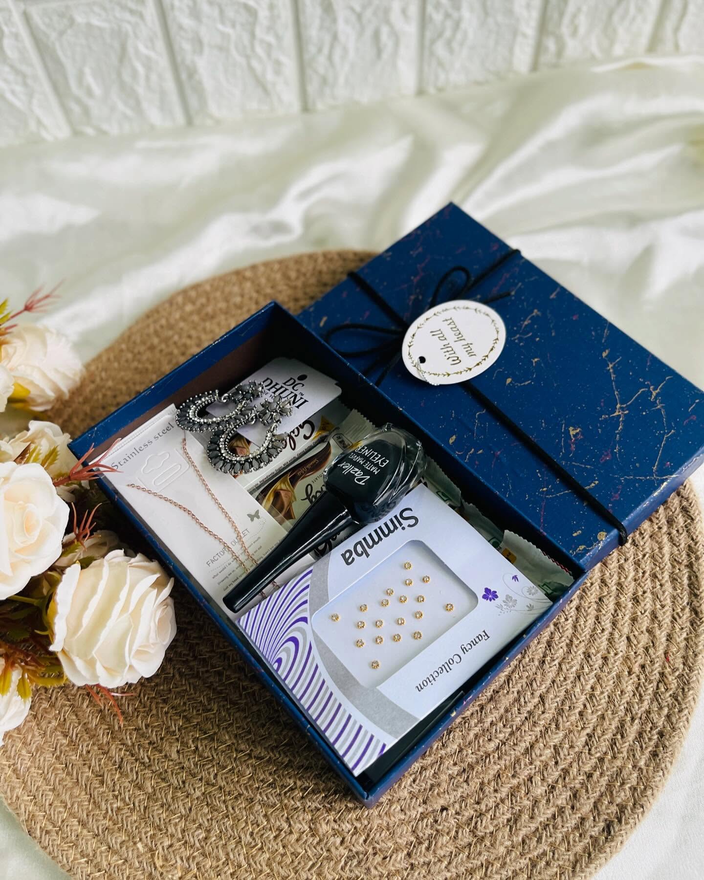 A navy gift box filled with statement earrings, a dainty necklace, nail polish and sweet treats, tied with a heartfelt tag.