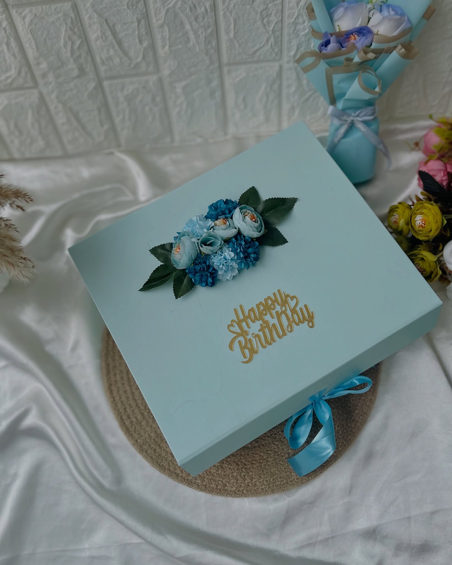 A soft teal gift box topped with blue and teal florals and a gold "Happy Birthday" topper, paired with a matching flower bouquet.