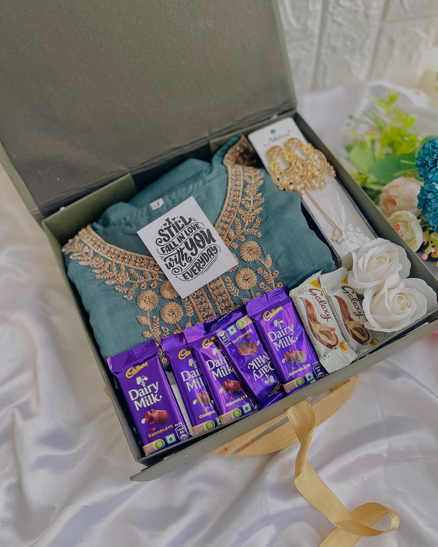 An open gift box featuring an embroidered teal kurta, gold-tone jewelry, chocolates and roses.