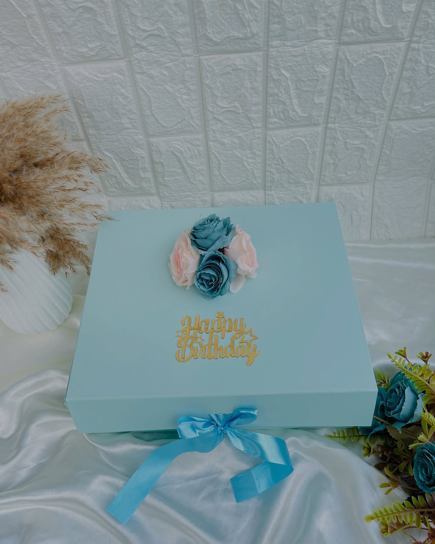 A pastel teal gift box topped with a gold 'Happy Birthday' script and a cluster of teal and blush roses.