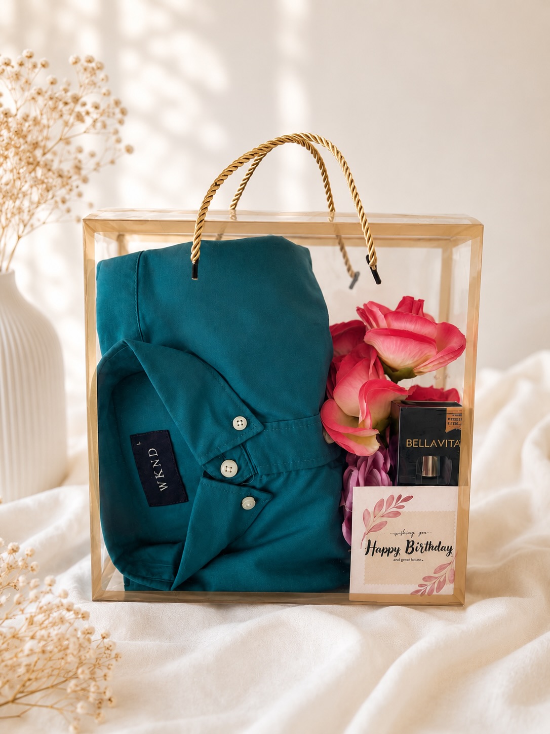 A clear gift bag styled with a teal shirt, fresh pink roses, a Bellavita perfume and a birthday message card.