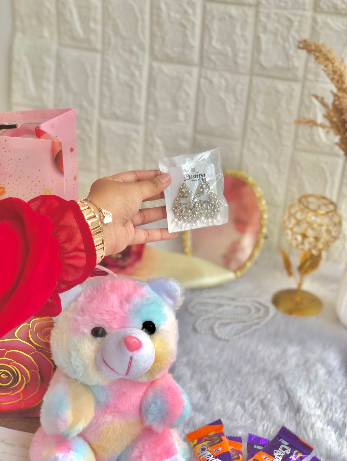 A playful birthday hamper pairing a rainbow teddy bear, sparkling earrings, and mini chocolates in a red gift box.