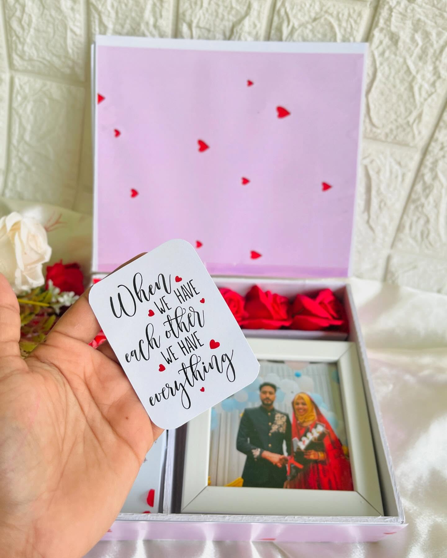 A pink heart-printed gift box holding fresh red roses, a photo frame, and a ring-bound Valentine's photo flip book.