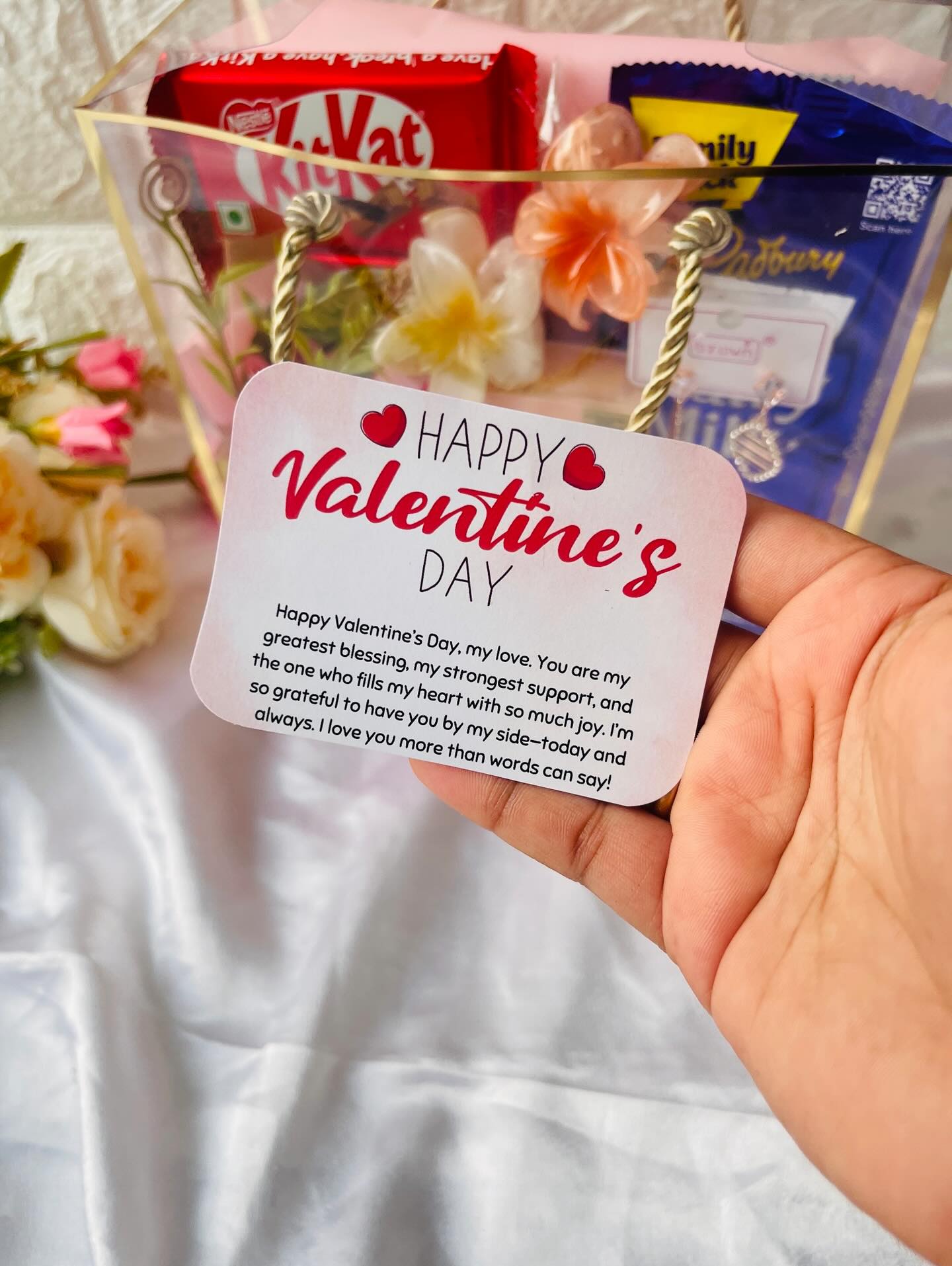 A romantic clear gift box with KitKat and Dairy Milk chocolates, a Bellavita perfume, drop earrings, and a rose bouquet.