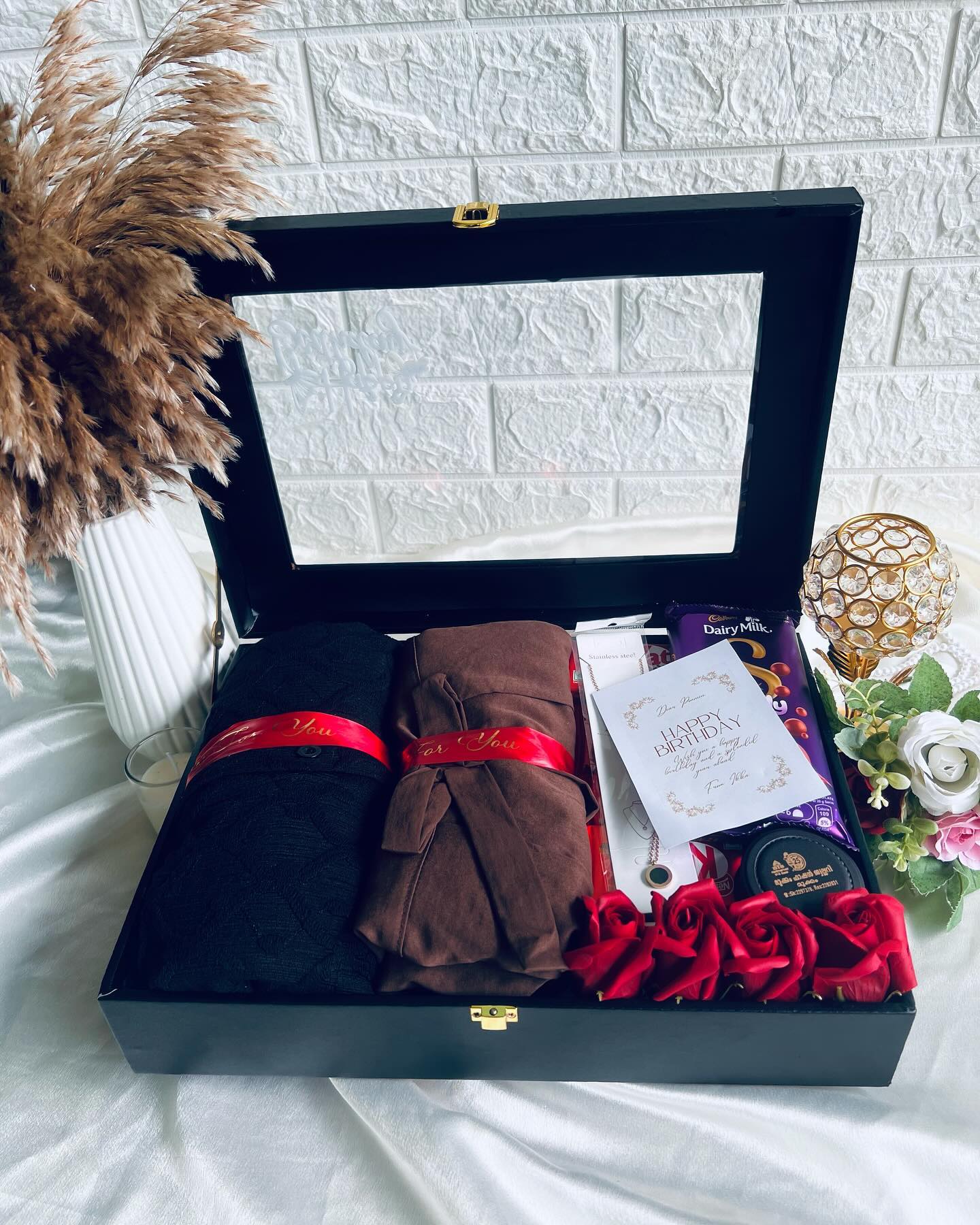 A glass-lidded birthday trunk with two ribbon-tied fabric wraps, jewellery, chocolate, and a personalized card and roses.