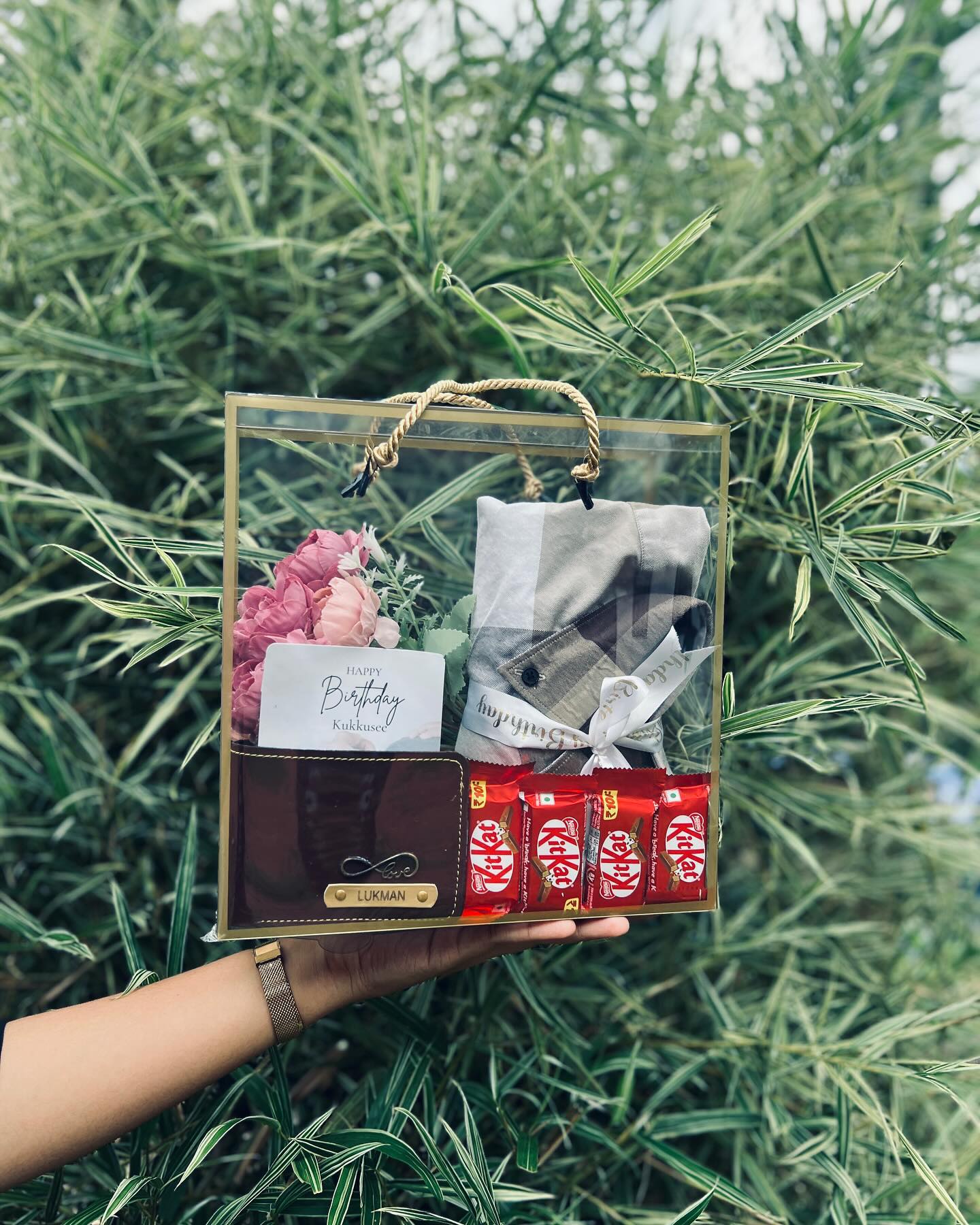 A clear-top gift box featuring a personalized monogrammed wallet, a checked shirt, fresh flowers and KitKat bars for his birthday.
