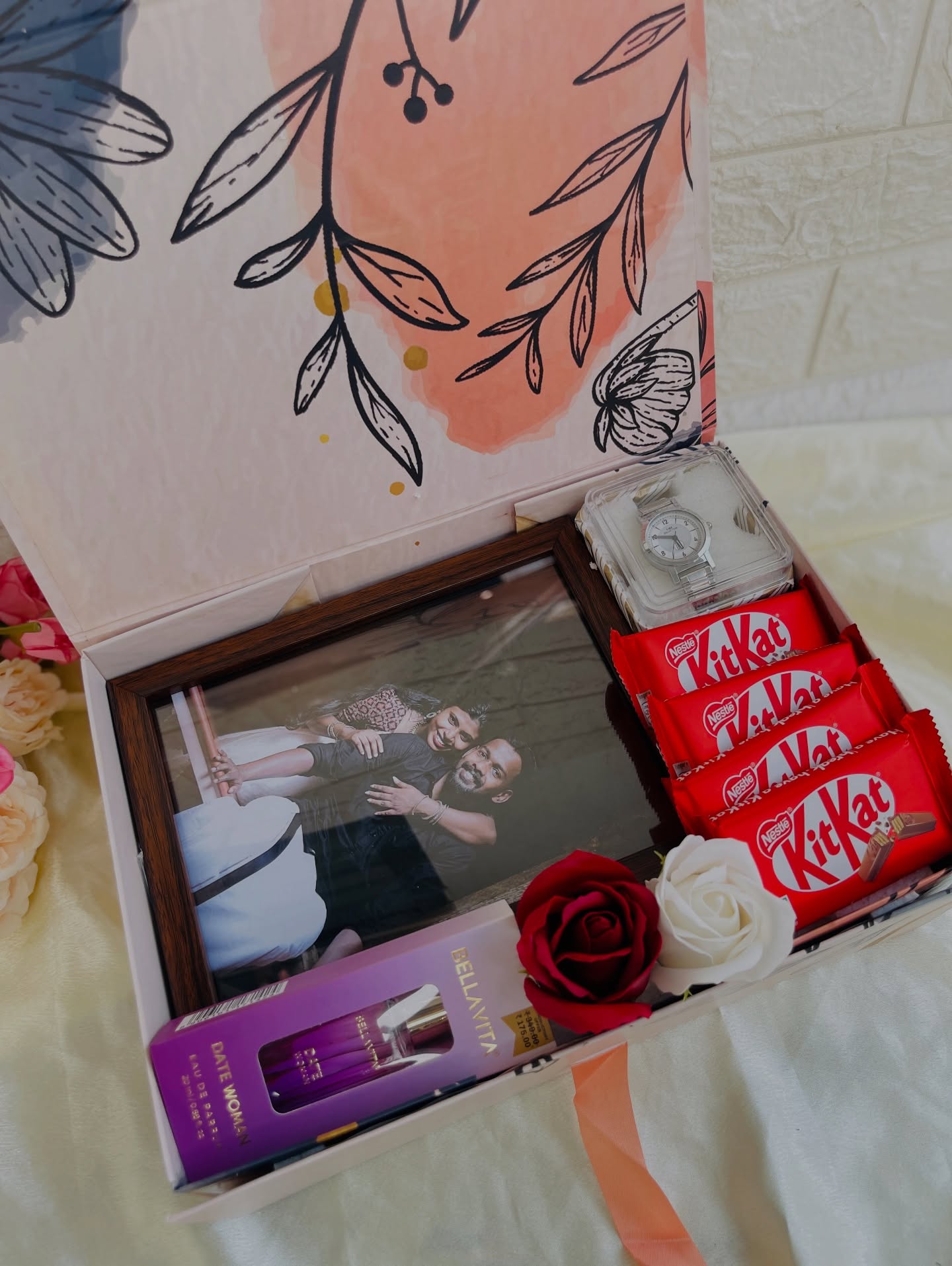 A floral gift box pairing a wristwatch, a fragrance, a couple photo frame, and KitKat bars.