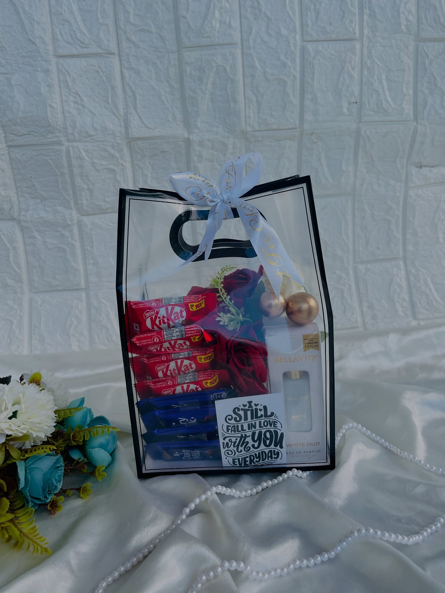 A clear gift bag filled with KitKat, Dairy Milk chocolate, a Bellavita White Oud perfume and a velvet rose.